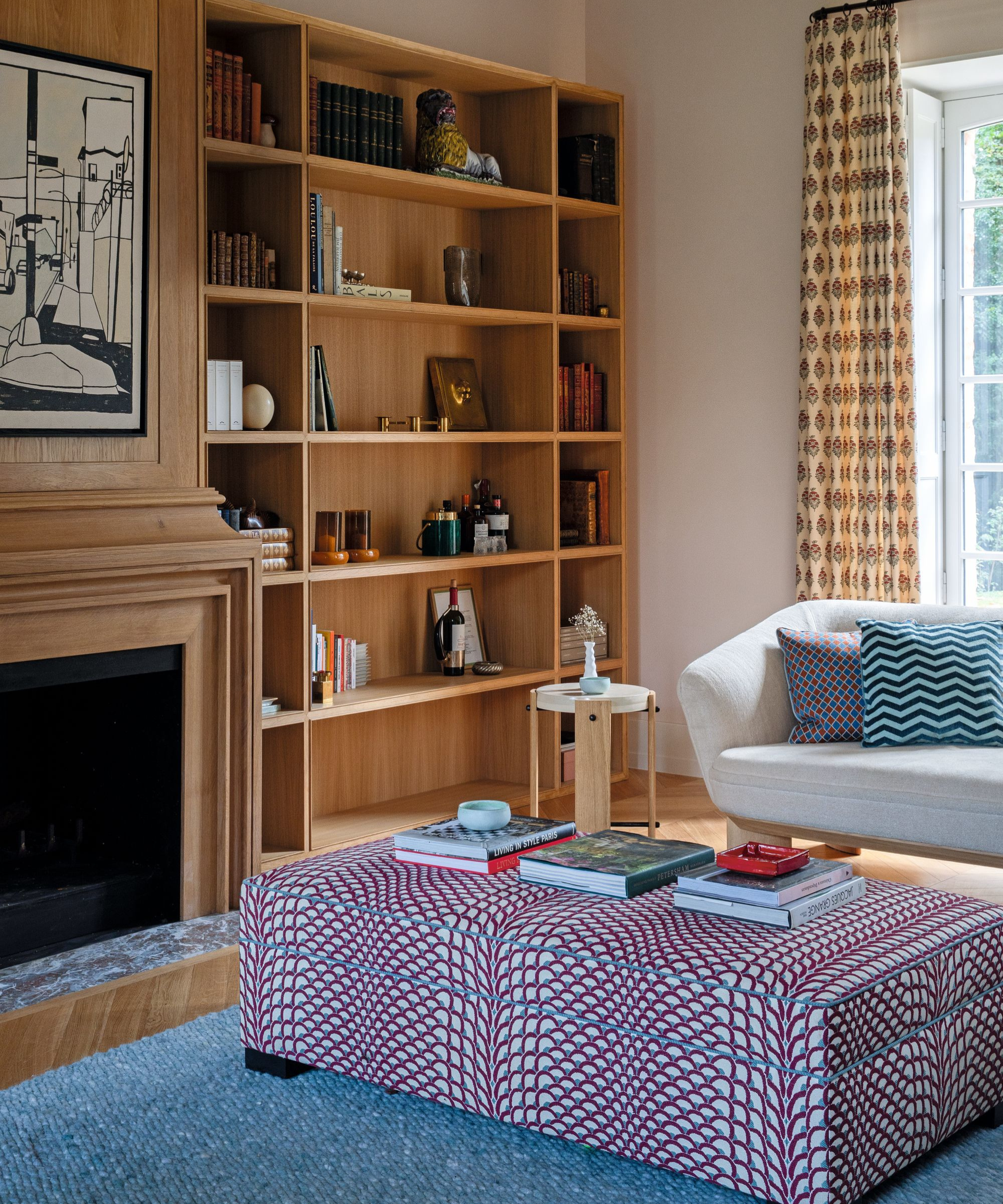 a living room with build in shelves with decor on them, a red and blue patterned ottoman, a blue rug, and books on top of the ottoman
