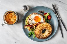 A cup of coffee and a bagel sandwich with avocado, fried egg and side salad on a white marble background, alongside a salt pot and knife and fork.