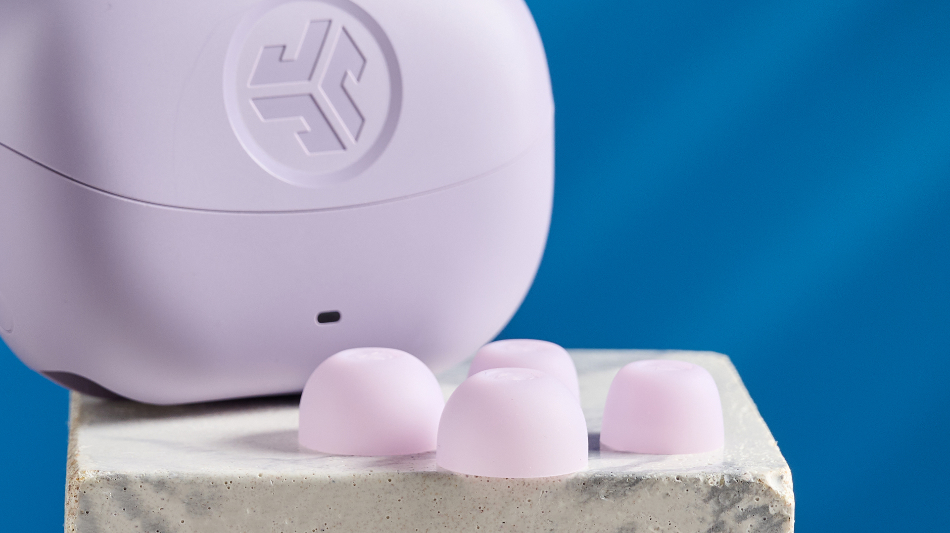 the jlab go pods anc earbuds in lilac with a lilac charging case and built in usb-c cable against a blue background on a marble stand