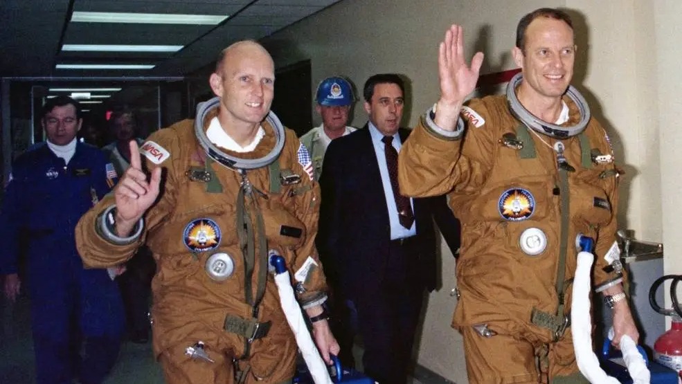 two astronauts in spacesuits waving in a hallway, with a group of people walking behind them