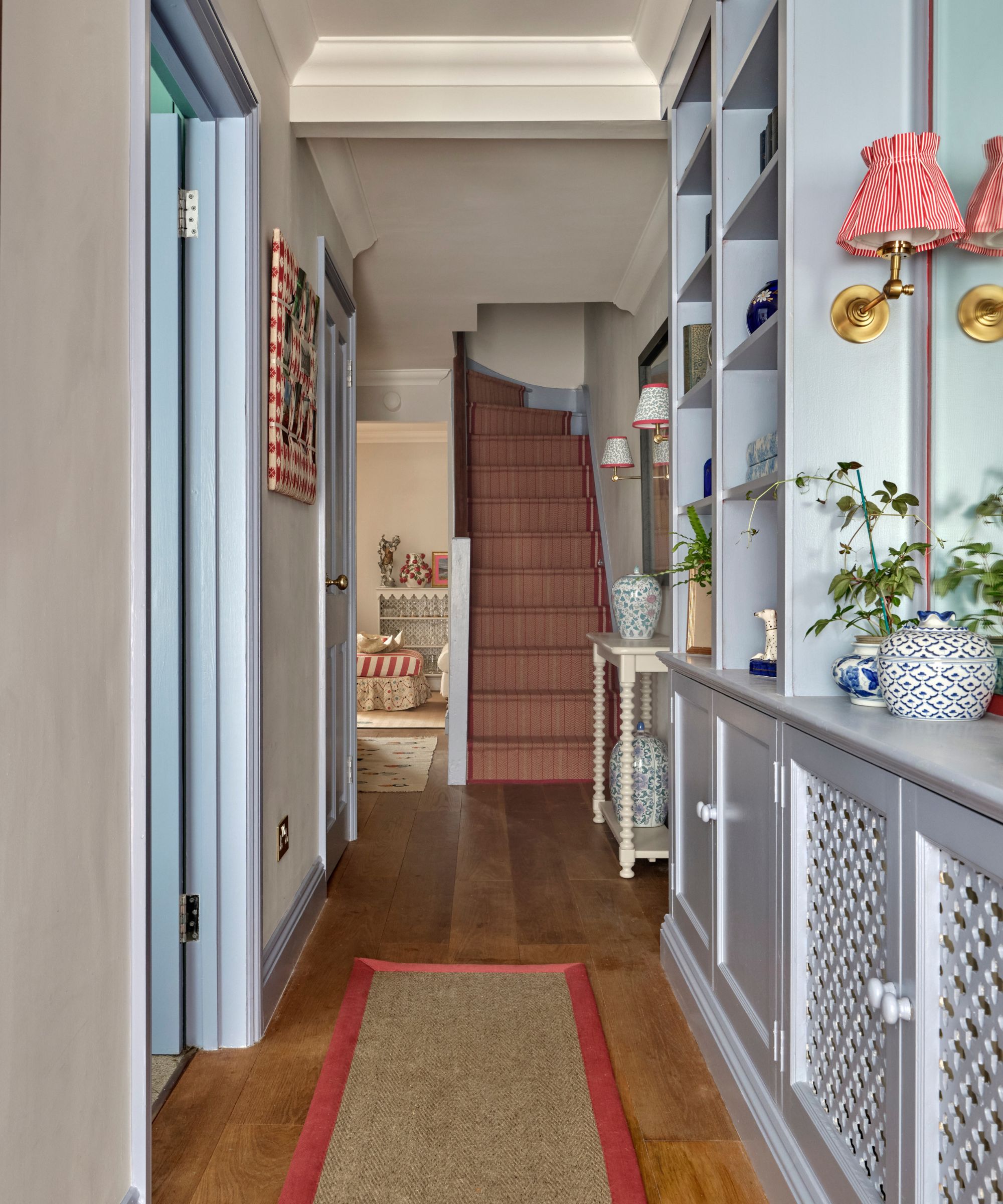 A narrow hallway featuring light blue built-in cabinetry and door frames, a natural fiber runner with a red border, and a staircase with a red striped carpet.