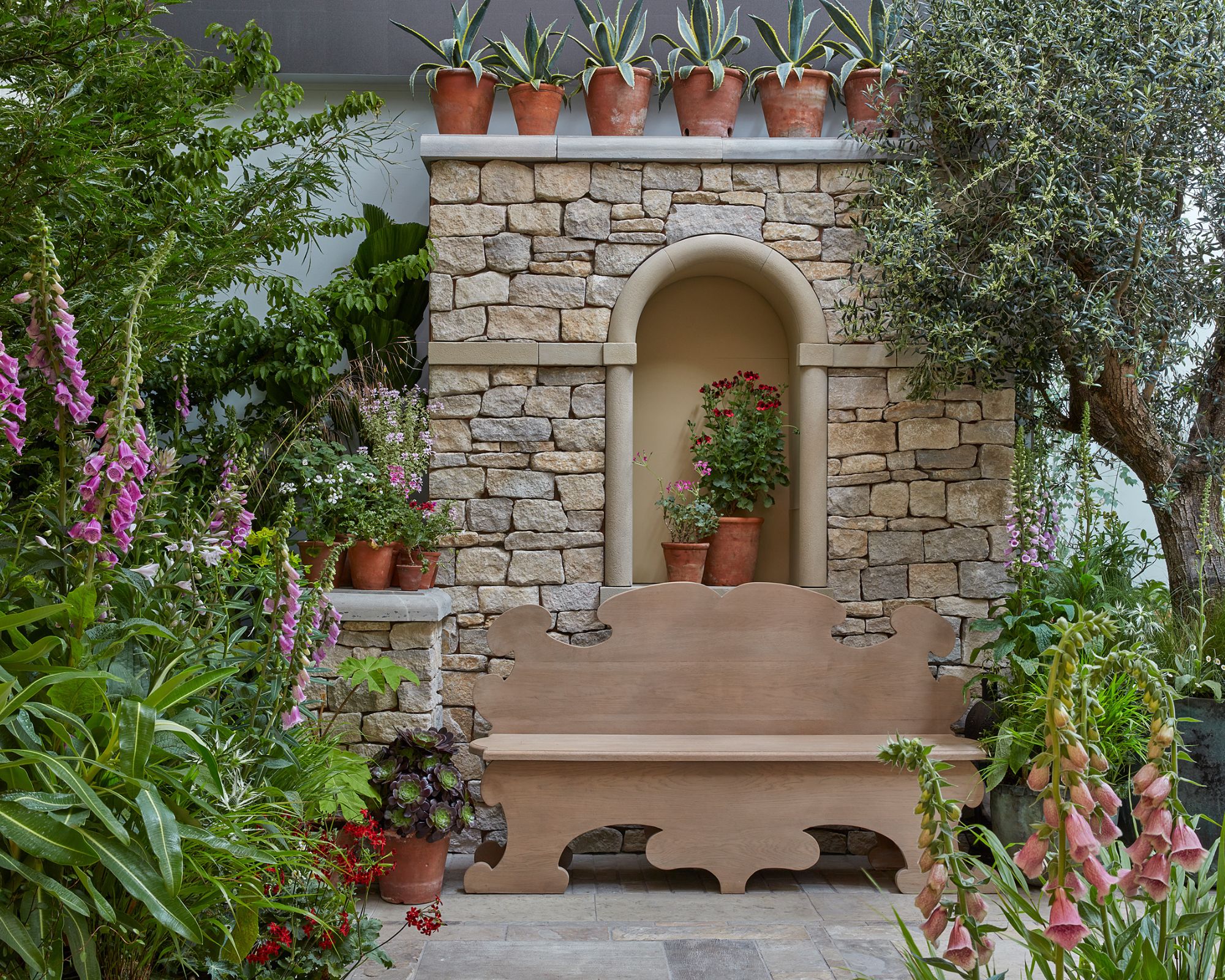 A stone wall in a garden with foxgloves either side and a wooden bench