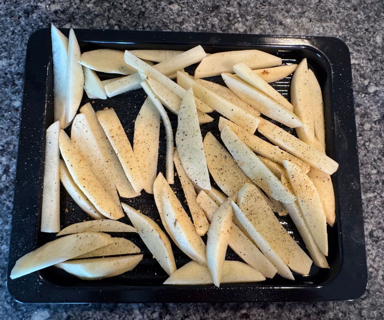 A black baking tray of raw potato fry slices