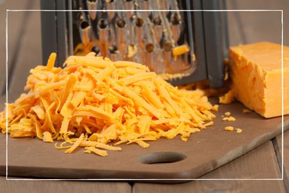 Grated cheese on a wooden board next to block of cheese with grater in the background