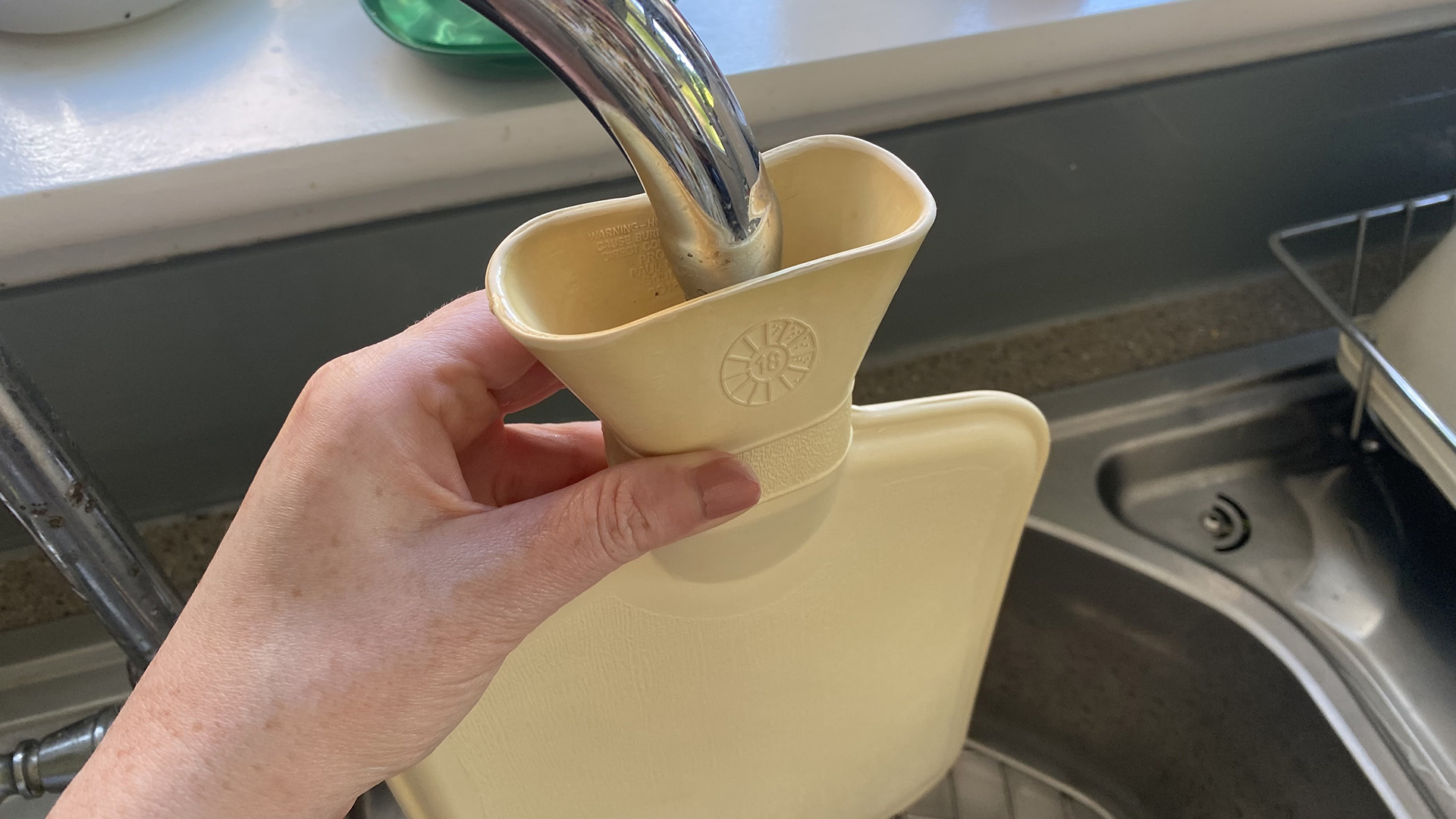 A close up of a hand holding the lip of a hot water bottle under a faucet as the water bottle is filled with cold water