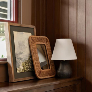 A small black lamp with a white shade sitting on a window sill in front of a small mirror and picture frame.