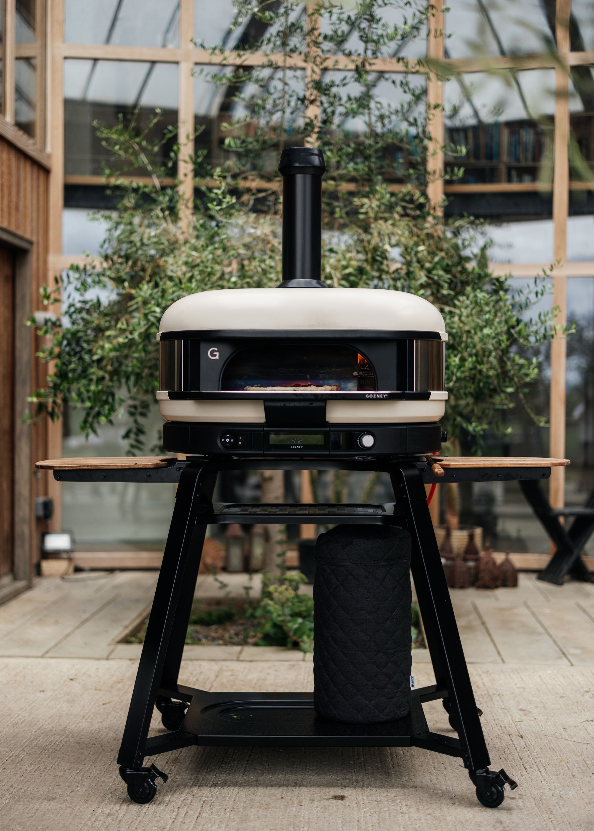 Gozney's Dome XL Gen 2 pizza oven in the bone colorway in an outdoor area with a backdrop of wood-framed glass panels and foliage