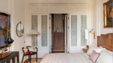 A calming bedroom with traditional wainscoting paneled doors leading to a wardrobe, framed art, floor lamps, a sideboard with a lamp and a rug