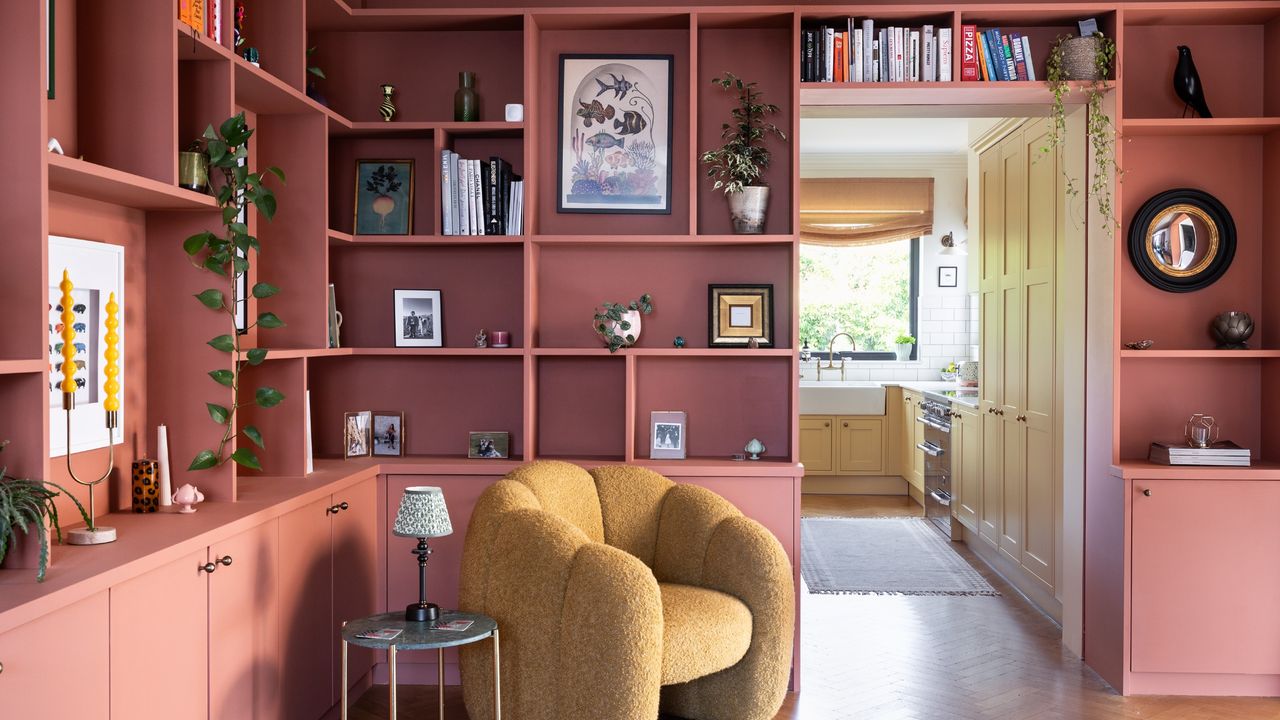 A built-in bookcase with bottom doors painted in a terracotta shade with a yellow boucle accent chair in front of it