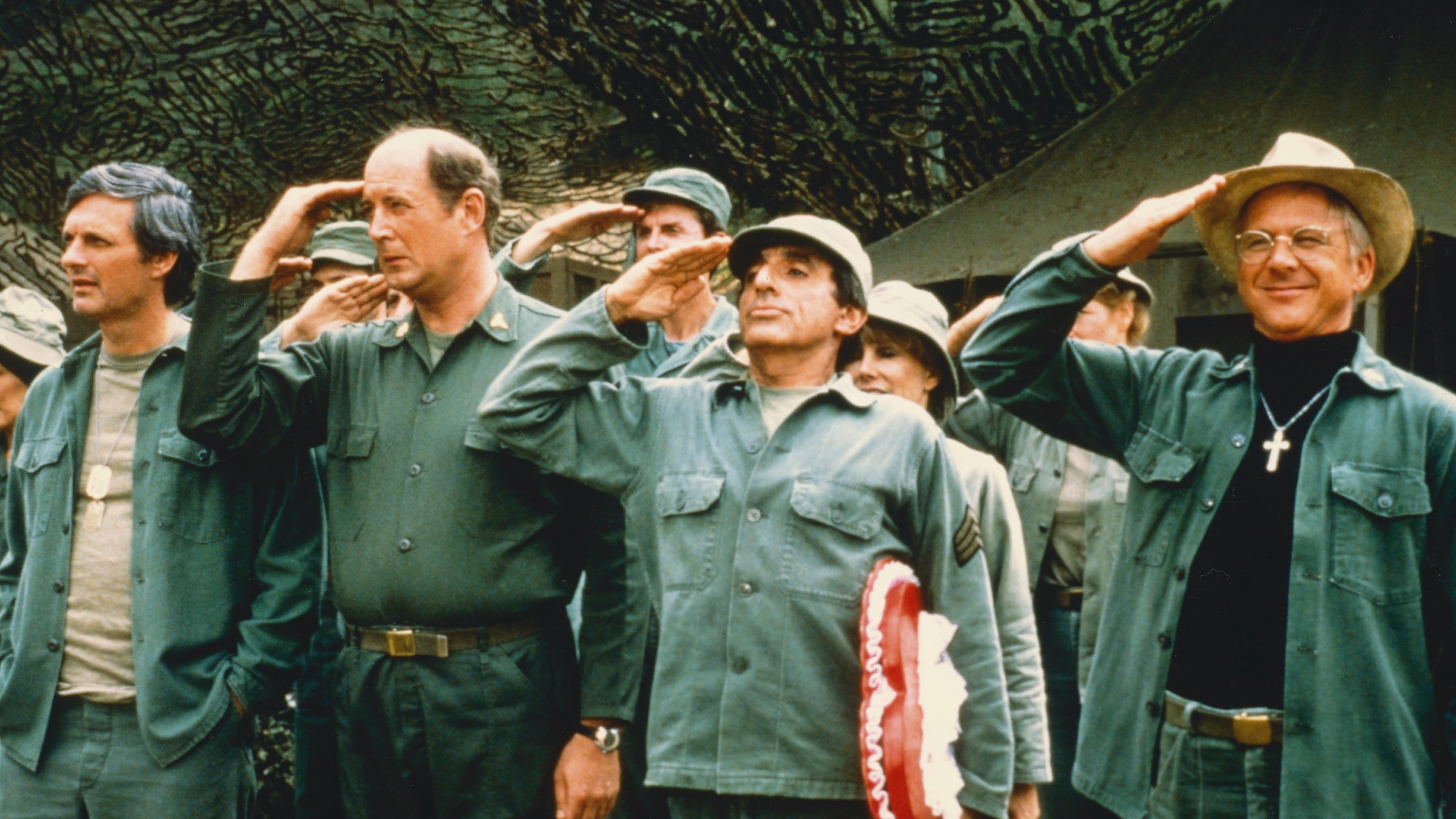 still from the TV show MASH, with four main characters standing in a line in fatigues giving a salute