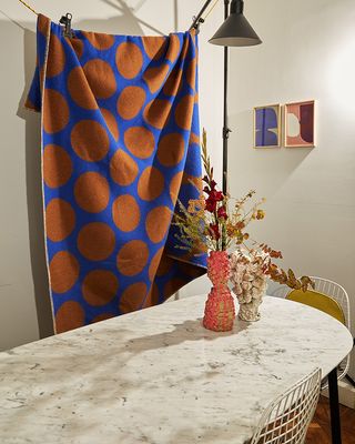 A room with a marble effect dining table, a vase with flowers and foliage, white chairs, a black lamp, framed wall art and a blue Colville woolen blanket with brown dots hanging out on a line
