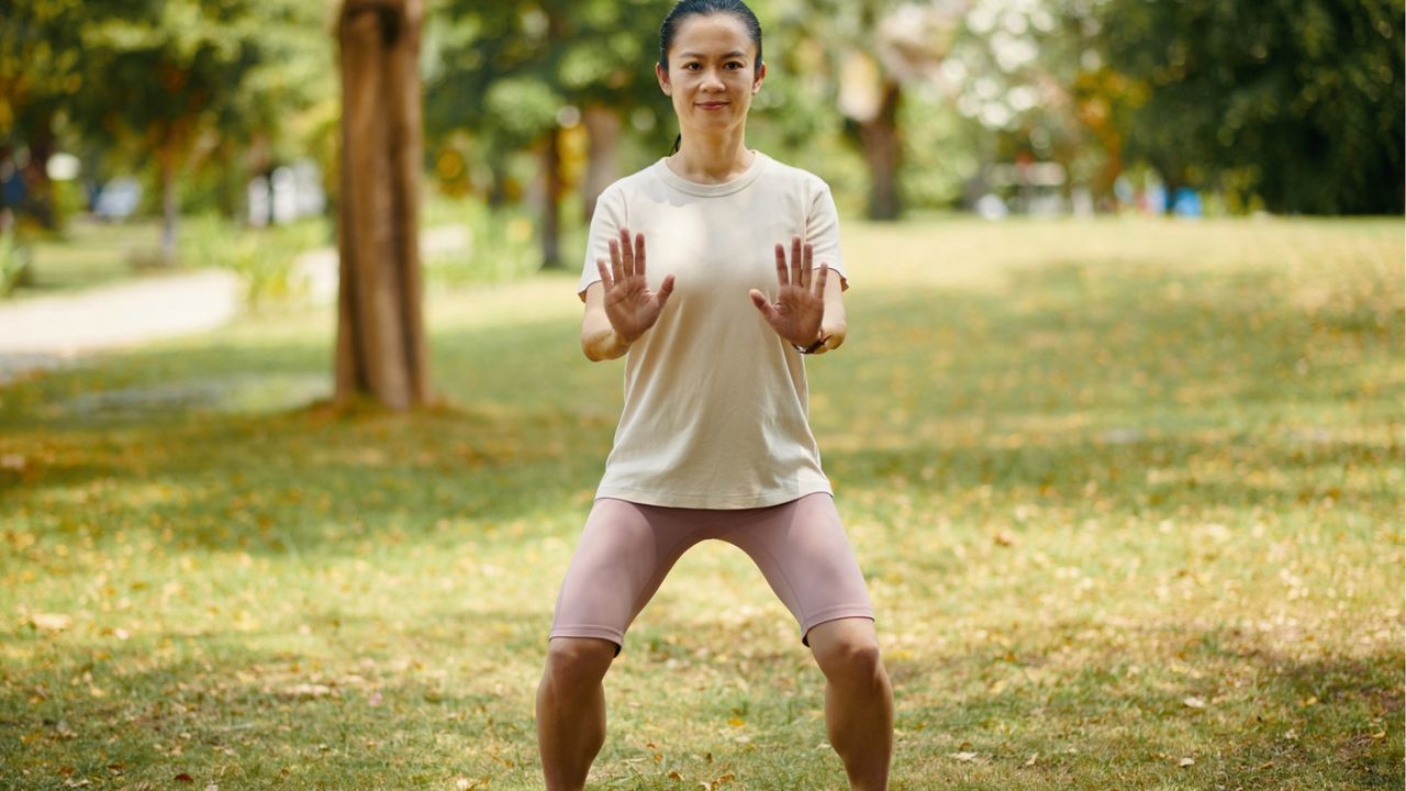 A woman practices Tai Chi in a park. She is standing with her legs gently bent, her arms straight and her palms facing forward. Behind her we see trees and grass.