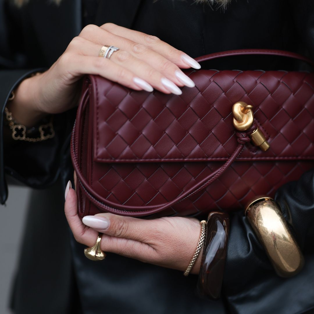 Hard BIAB Olga Liber with long, softly waved blonde hair worn in a center part, seen wearing a black satin lace camisole paired with loose charcoal silk trousers and an oversized black leather blazer. She completes the look with a gold-buckled YSL belt, oversized Prada shades, and a burgundy woven leather clutch