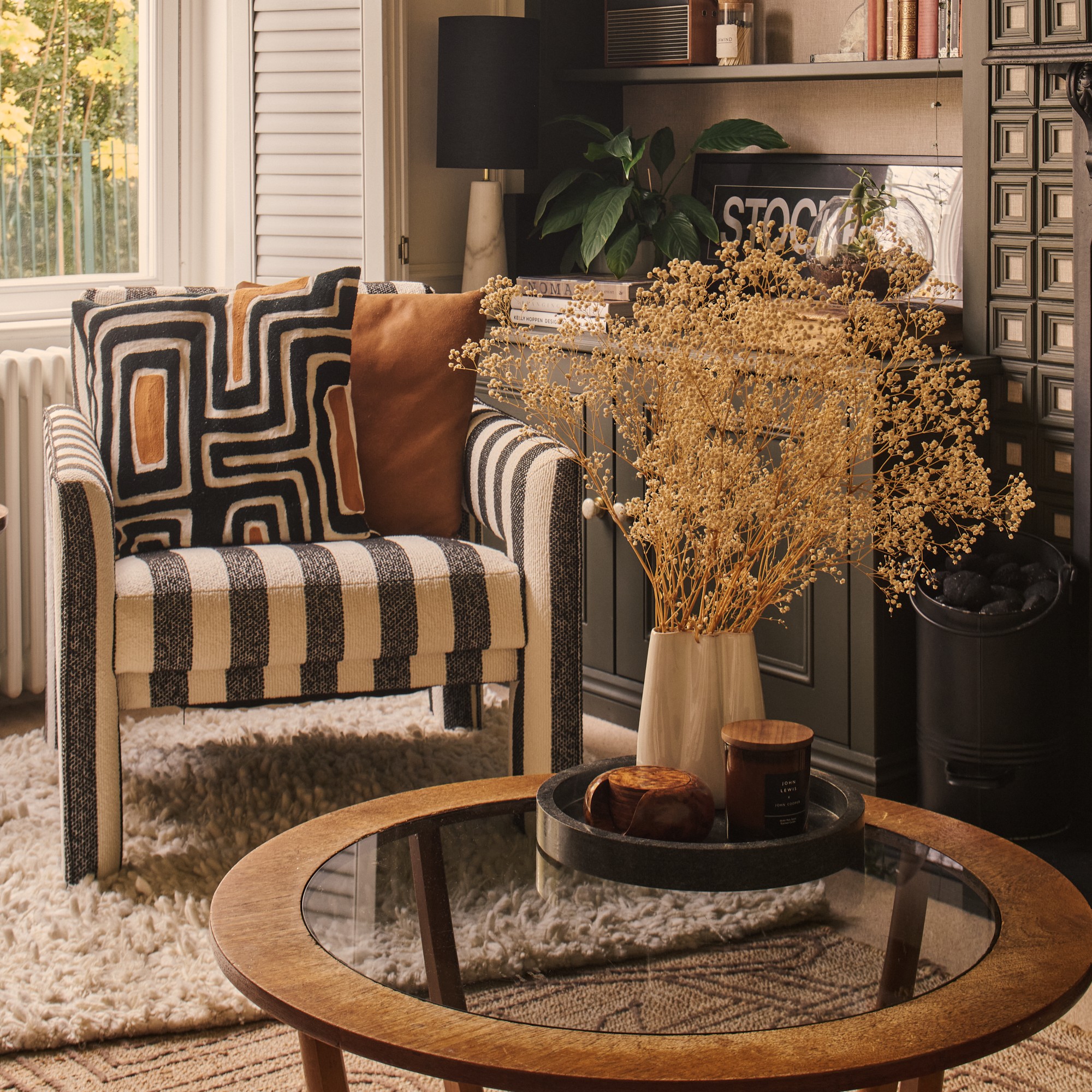 An earthy living room with a striped accent chair and a round glass and wood coffee table displaying a vase of dried flowers with a scented candle