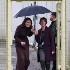 Kate Middleton walking through prison gates ducking under an umbrella that is being held by a woman in a blazer making a surprised expression