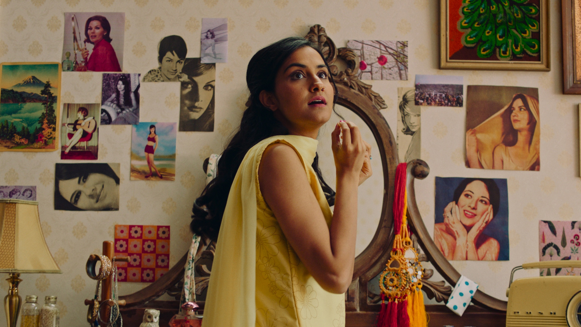 amrit kaur as young miriam in a yellow dress looking at a wall of old photos in the movie the queen of my dreams