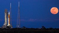 An orange full moon shines to the right of a large rocket standing on a launch pad in the darkening blue evening sky.