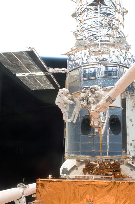 A metallic-wrapped spacecraft is seen close up. Astronauts are servicing it.