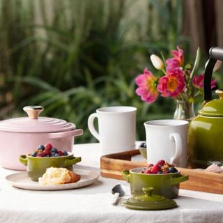 Let Creuset mini round cocotte, two green designs on table outside, filled with fruit, mugs and pink dish in background