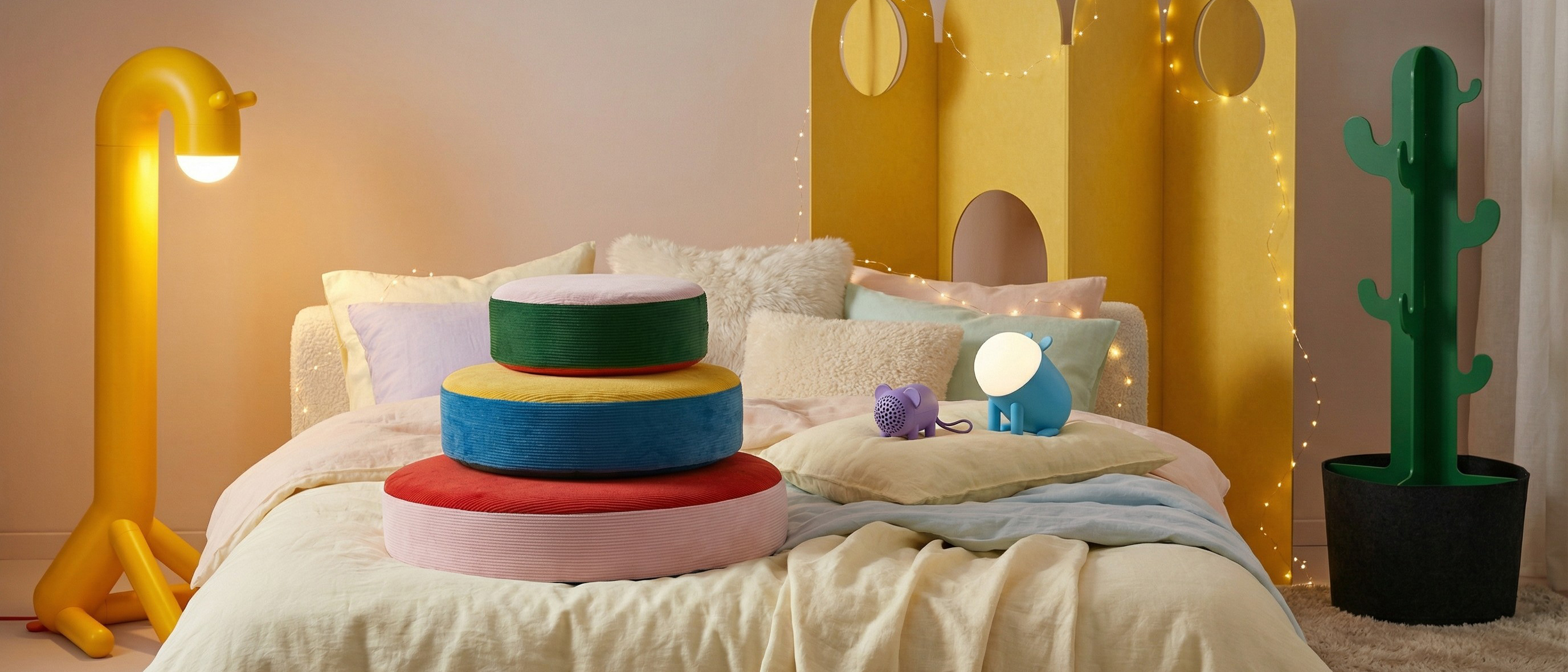 IKEA Grejsimojs homeware products including a dog-shaped lamp, giraffe lamp, cactus coat hook and floor cushions on a bed, with twinkle lights in the background