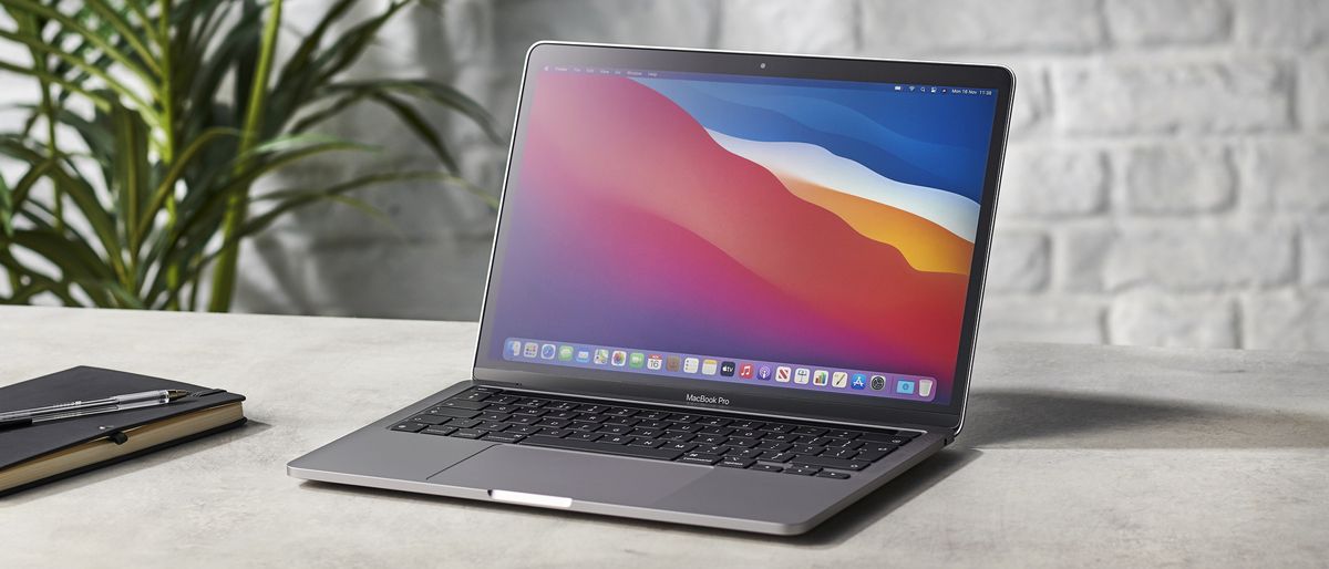 The Apple MacBook Pro 13-inch (M1, 2020) on a desk next to a notebook in front of a white brick wall