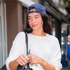 Dua Lipa exiting Electric Lady in NYC wearing a backwards baseball cap, white long-sleeve T-shirt, cut-off denim shorts, and a black Chanel Maxi Flap bag and burgundy Chanel Coco Beach ballet flats 