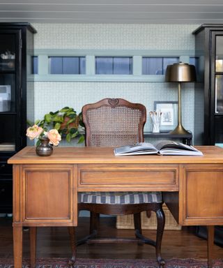 office with patterned blue wallpaper, black cabinets, antique wooden desk and cane back chairs