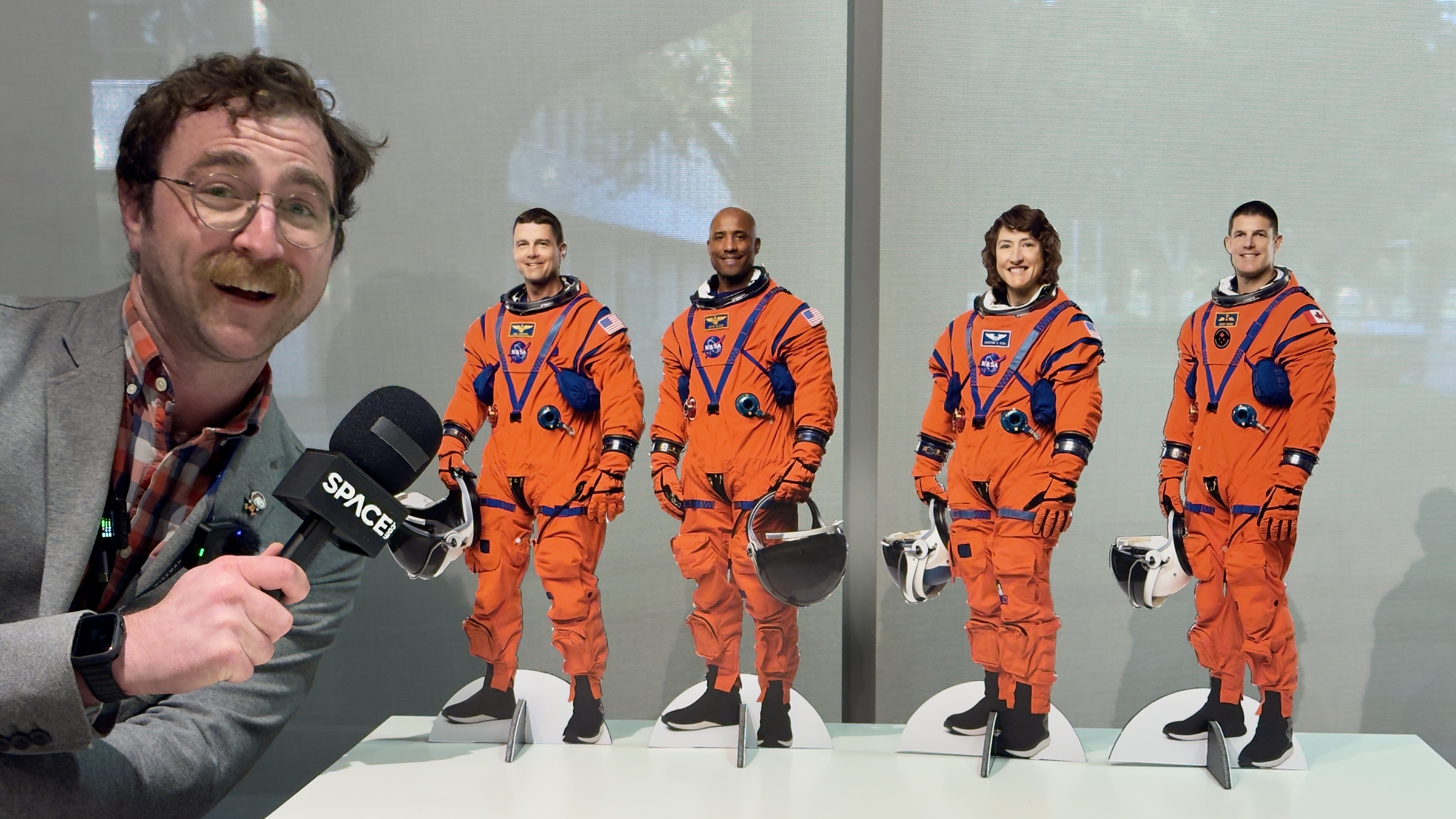 A giant man with a microphone leans over four tiny (cardboard cutout) people.