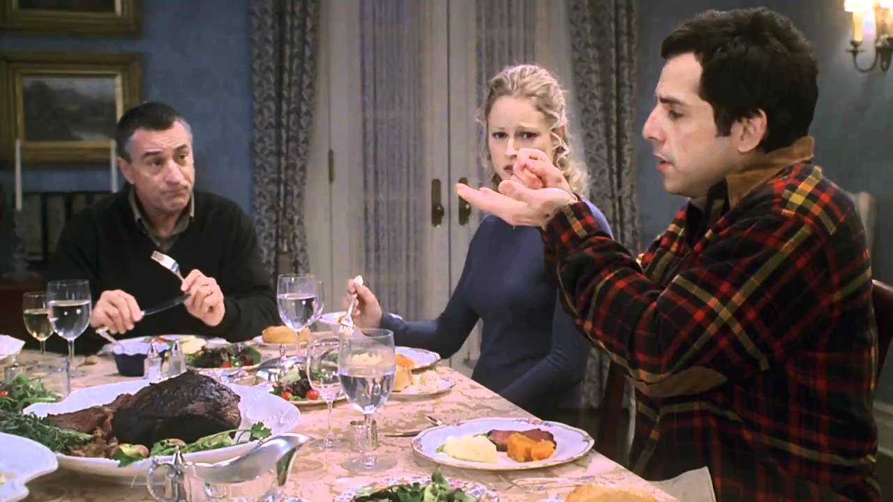 A still from Meet the Parents in which Ben Stiller is holding something at the dinner table and Robert DeNiro's character is staring at him.