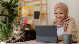 a smiling woman studies on a laptop while a large tabby cat lies next to her