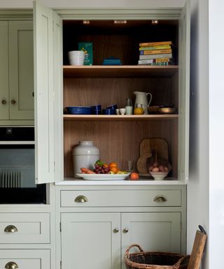 A sage green kitchen cupboard with wooden-lined open shelving. The top shelves display cookbooks, blue ceramic dishes, and a few small decor items. The countertop holds a large ceramic jar, a fruit bowl with fresh produce, and a wooden bowl with eggs. Below, closed cabinets with brass cup handles complement the rustic styling.