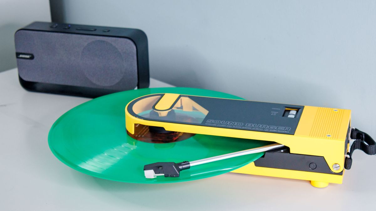 Yellow Audio Technica Sound Burger turntable on a white table top, playing a green record next to a black Bose SoundLink Home Bluetooth speaker