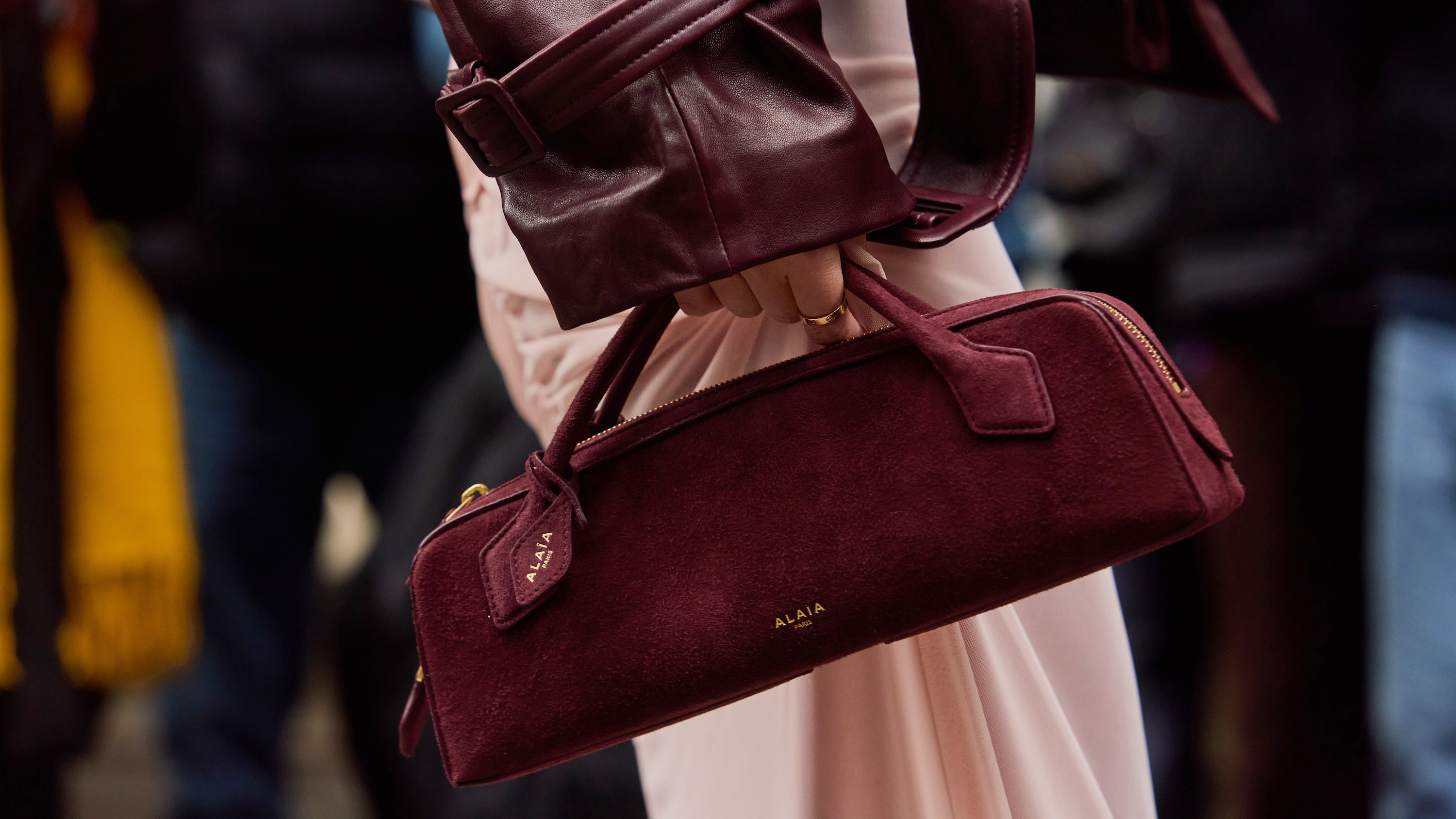 detail street style image of a person holding a burgundy alaia baguette bag with a burgundy leather sleeve jacket and light pink skirt