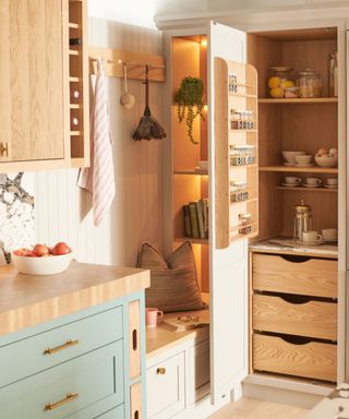 A cosy kitchen corner with a built-in pantry and bench seating. The pantry has a light wood interior with open shelves storing glass jars, cups, and a French press.The cabinetry is a mix of pale green and cream with brass hardware. Warm under-shelf lighting highlights the wood tones.