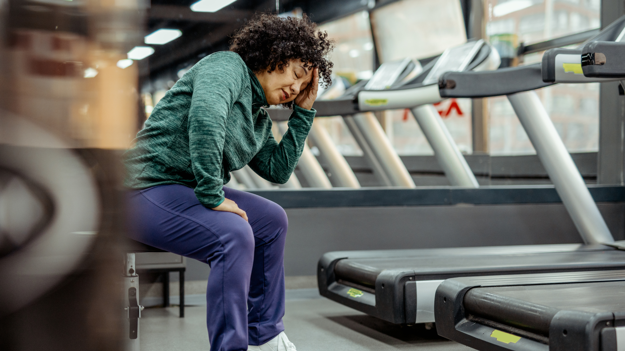 Woman sits in a gym with one hand on her forehead and her eyes closed