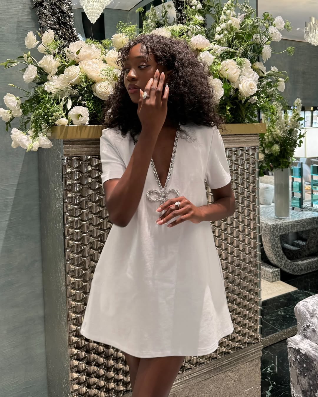 Emmanuelle Koffi wearing a white babydoll dress with a silver bow while attending a Graff high jewelry presentation