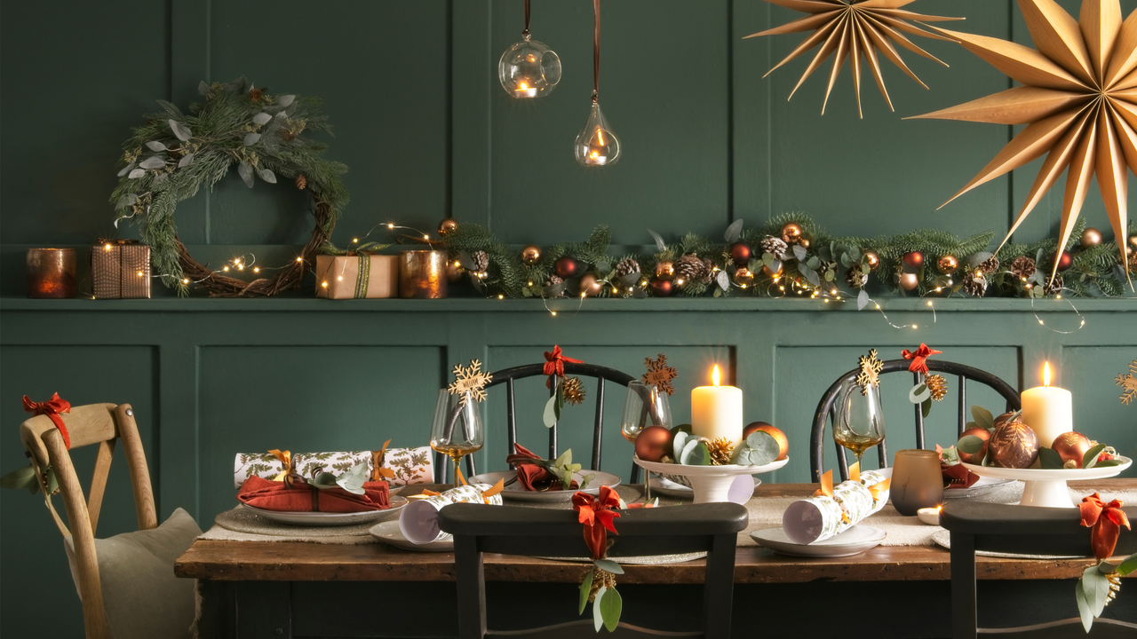 Table laid for a meal at Christmas with foliage and Christmas decoration above and Christmas decorations and lit candles in the background.