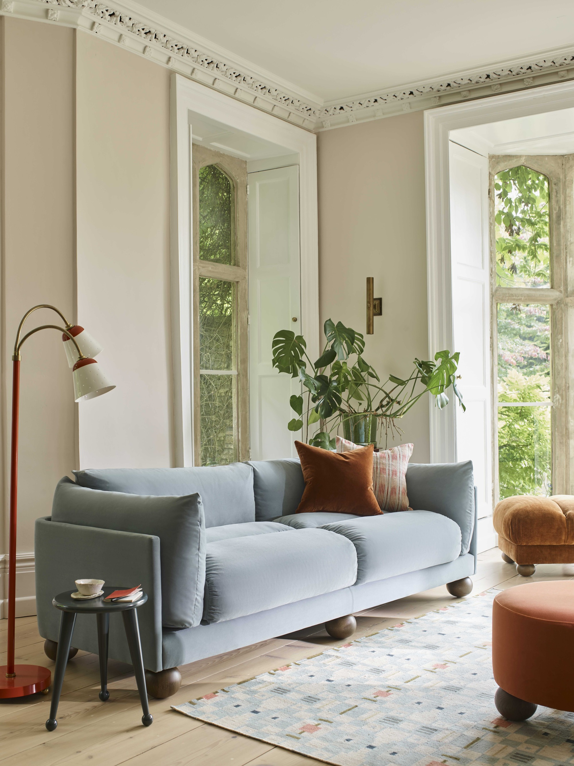 a light blue velvet sofa in a traditional living room with timber bun feet, blonde floorboards, pale pink walls with ornate cornice, a potted monstera plant and window behind the sofa, a side table and orange floor lamp, and blue and orange rug on the floor
