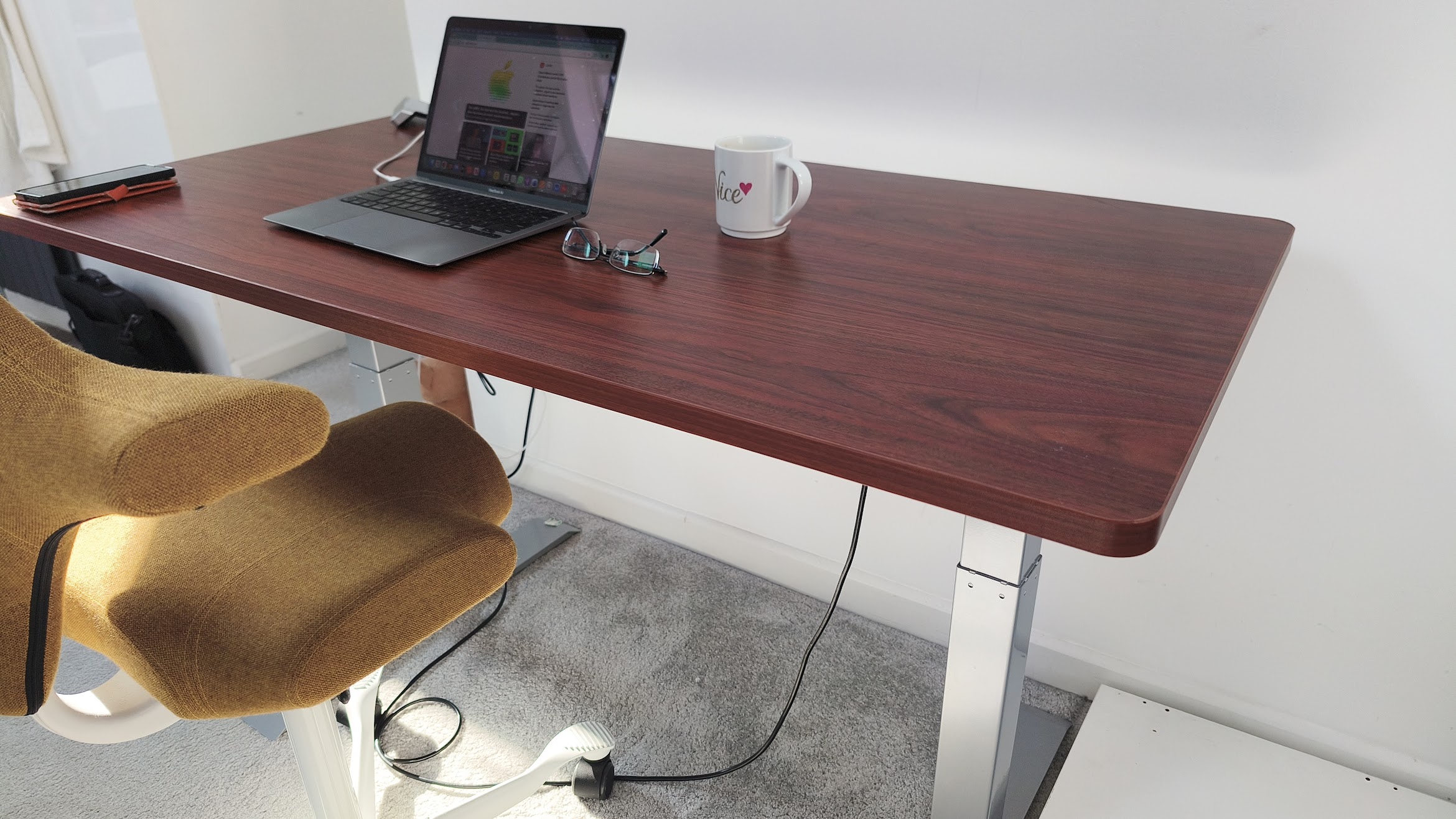 A wide shot of a mahogany-finish standing desk in a bright room, featuring a laptop, a white mug, and a pair of glasses on its surface.
