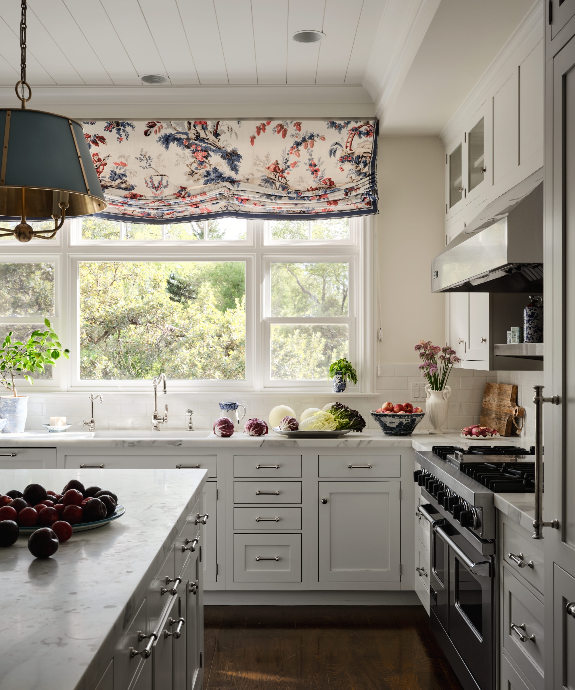White kitchen with marble counters, paneled cabinetry, a large garden window and a red-and-blue patterned Roman shade.