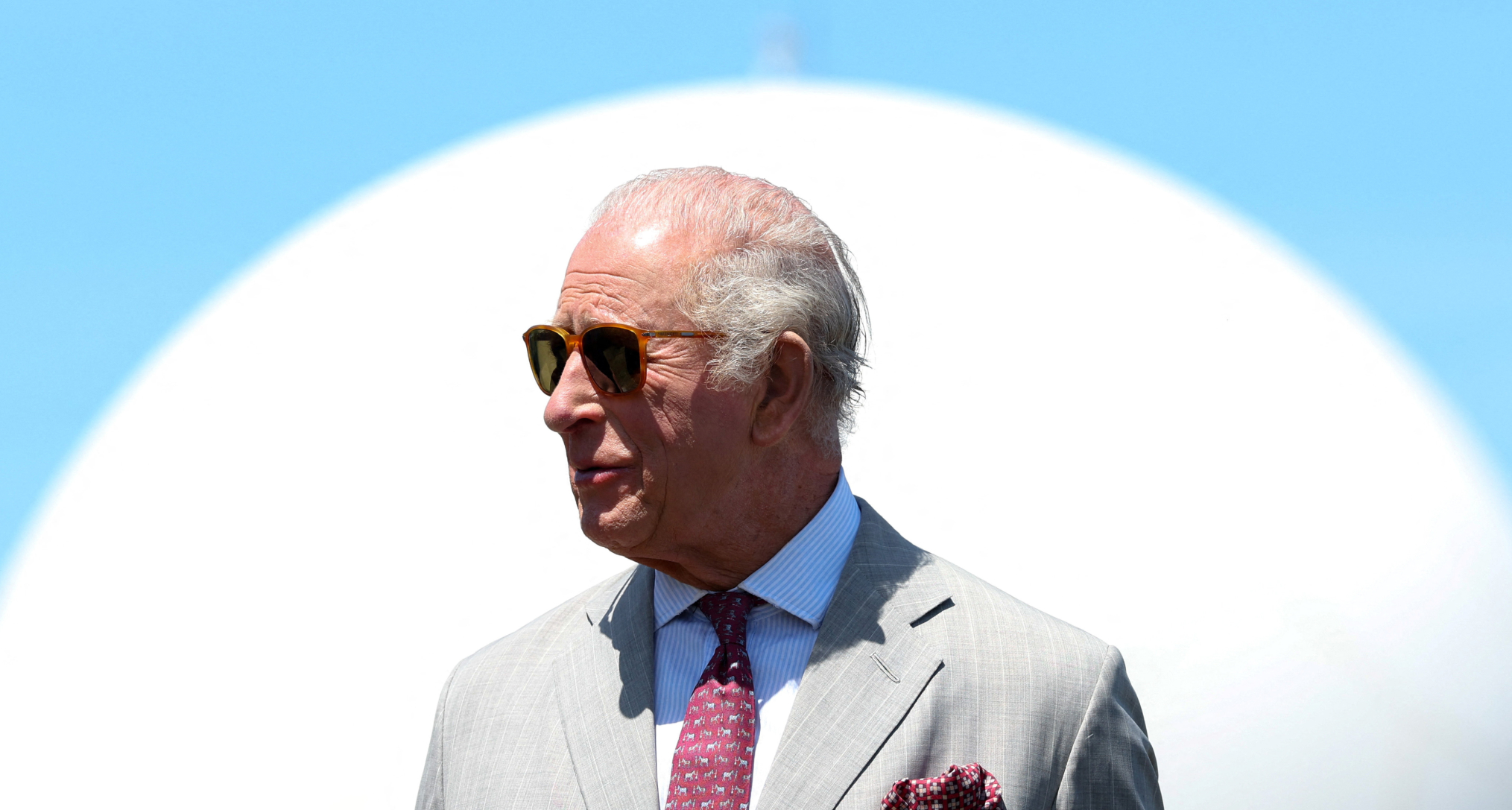 King Charles wearing sunglasses standing against a blue and white background