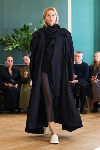 A photo from the Carven S/S 25 runway show of a model wearing a black maxi-length trench coat with a sheer skirt and cream slides.