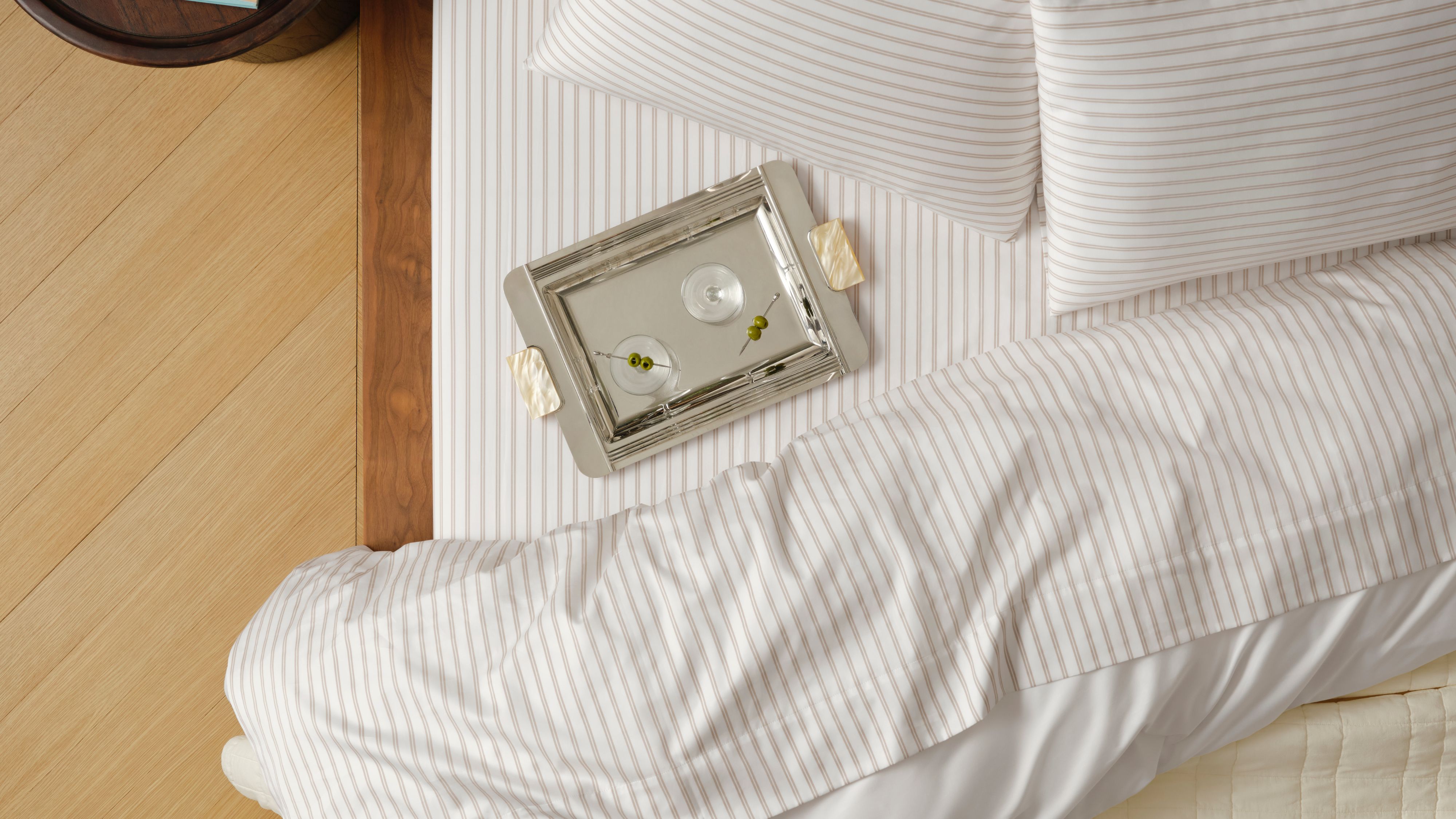 Brooklinen striped luxe sateen bedding and silver martini tray pictured from a bird's eye view inside of a neutral bedroom with hardwood floors