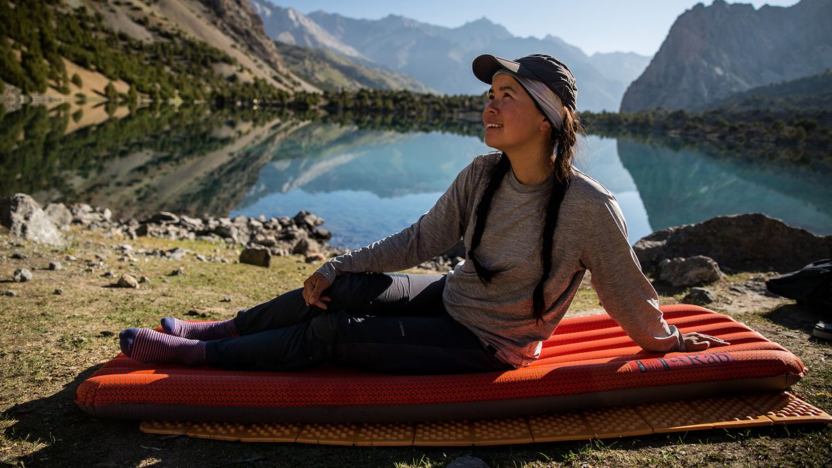 A woman sits back on an orange Rab Ionosphere 5.5 Sleep Mat, a lake behind her.