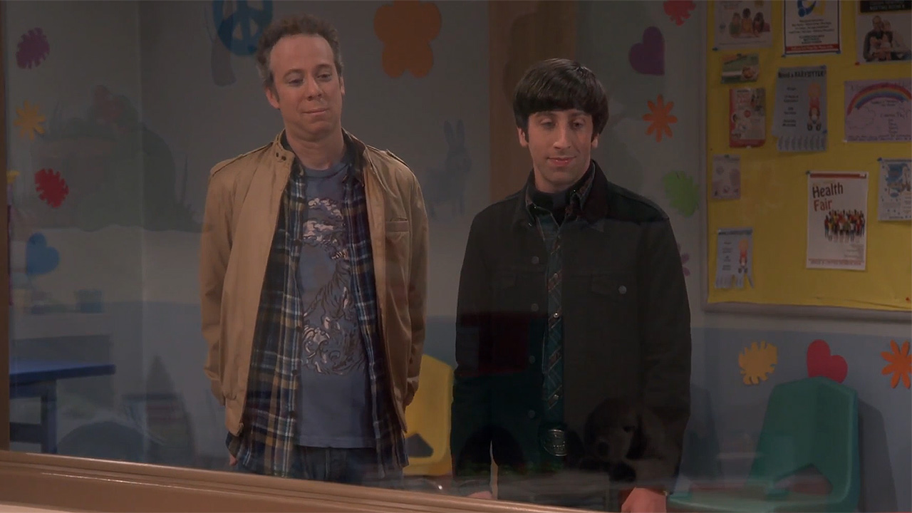 A screenshot of Howard and Stuart looking through a window into a daycare on TBBT. Howard has a dark jacket on and Stuart is wearing a camel coat.