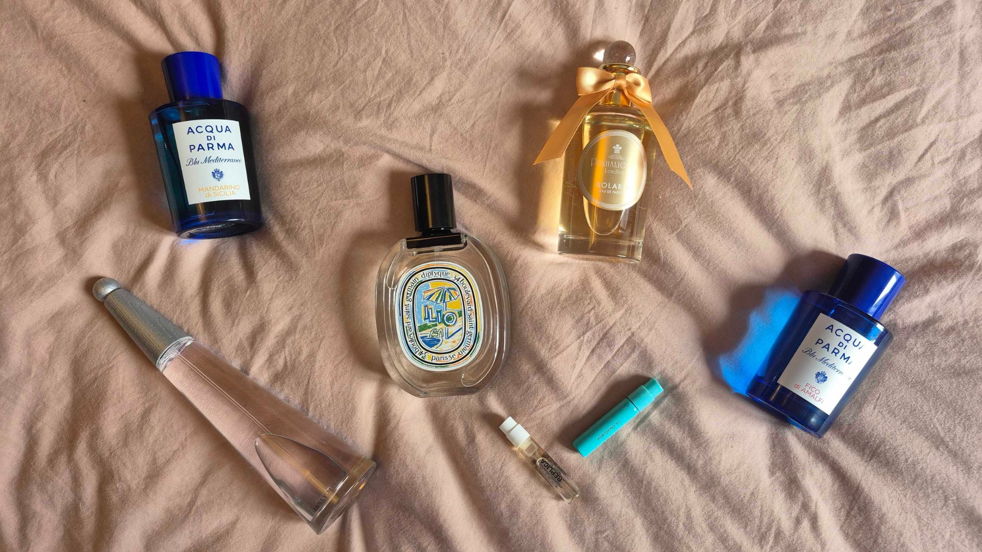 A selection of some of the 'Riviera perfumes' featured in this roundup, including two of Acqua Di Parma's Blue Mediterraneo scents, Issey Miyake's L'eau D'issey, Penhaligon's Solaris, Diptyque's Ilio and samples of Maison Margiela's Beach Walk and Victoria Beckham Beauty's Portofino '97, all laid out on a pink bedspread background