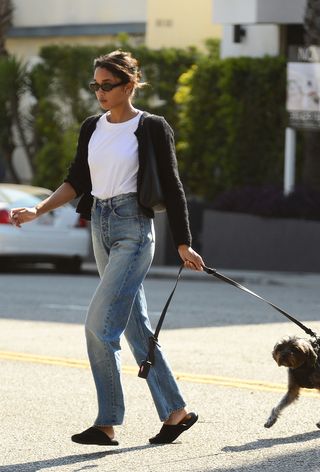 Amazon Prime dupe find of the celebrity-approved straight-leg jeans trend shown on a candid photo of Laura Harrier wearing black sunglasses, a black shoulder bag, a black cardigan, a white t-shirt, light wash jeans, and black mules