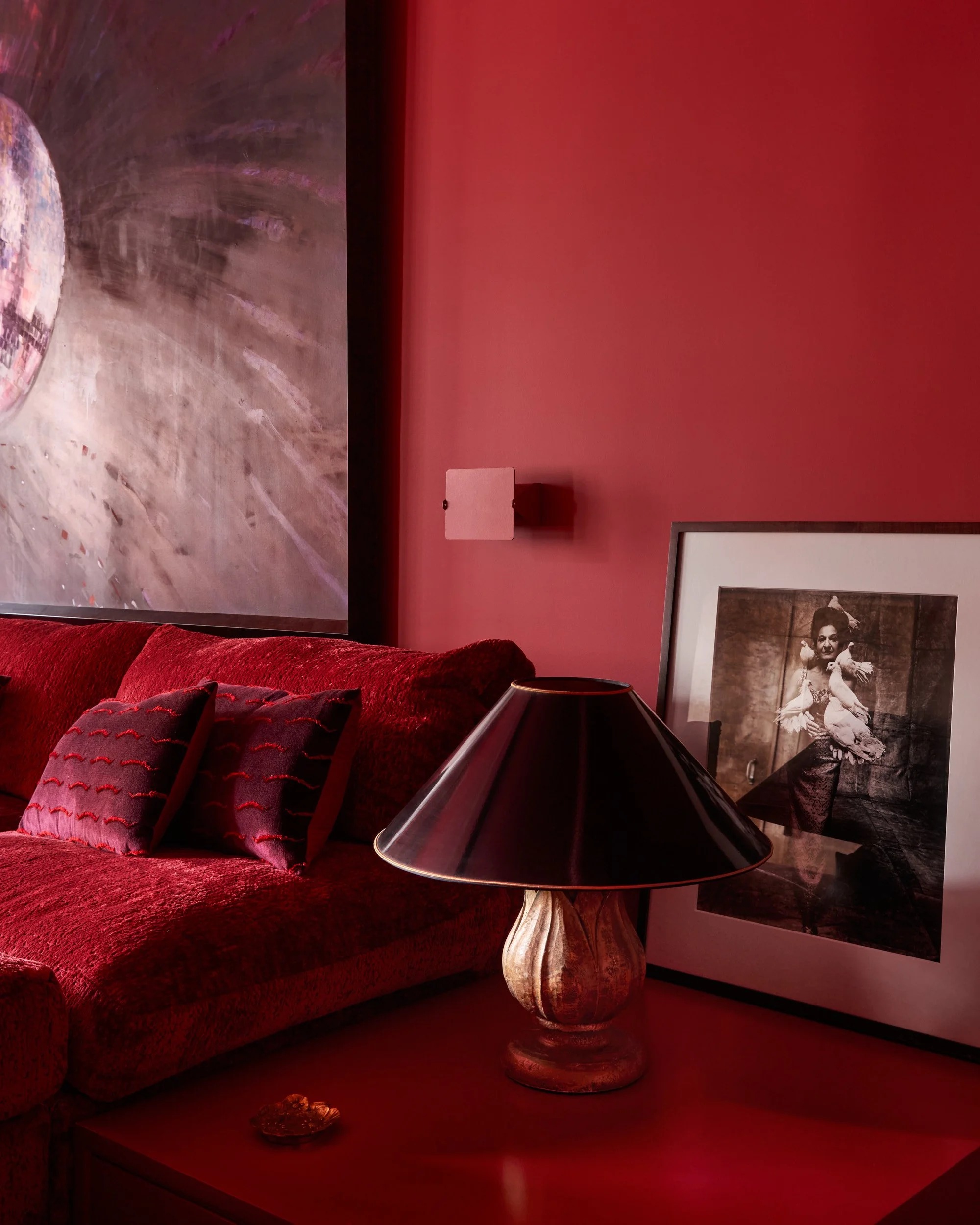 Image of a red room with the corner of a red sofa and a red side table in view. There is a black and gold lamp on the table and a picture frame.