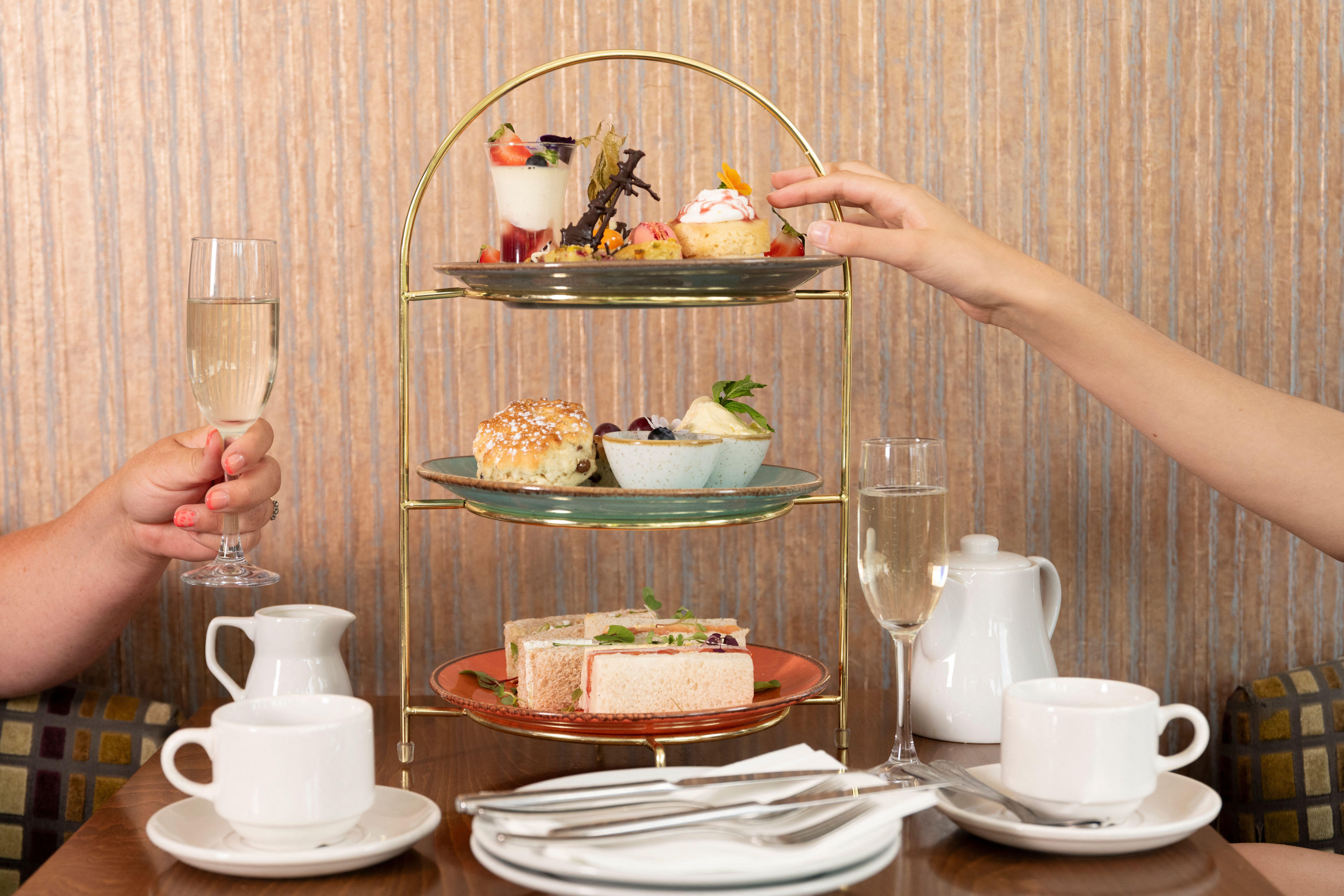 Two people enjoying afternoon tea, with cakes, sandwiches and champagne. Only their hands are visible