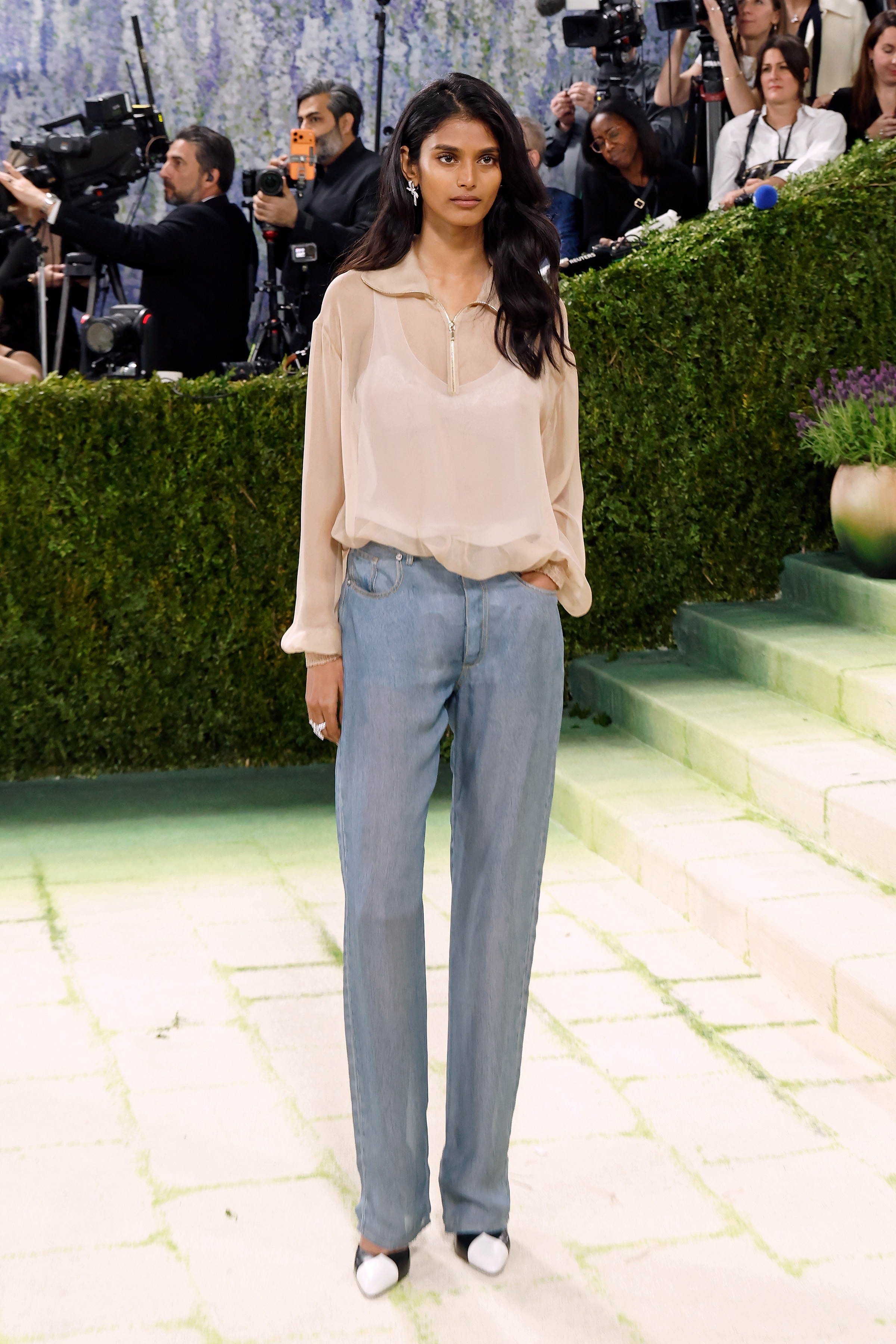 Bhavitha Mandava poses on the 2026 Met Gala carpet wearing a sheer beige zip-up blouse, loose light-wash trompe l&amp;rsquo;oeil denim trousers, and black-and-white cap-toe heels by Chanel